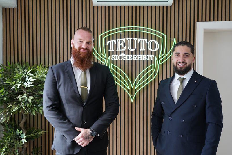 Jan Lockhorn und Omar Khattab im Anzug vor dem beleuchteten Logo der Teuto-Sicherheit
